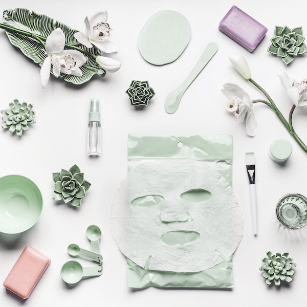 Green skin care cosmetic setting with orchid flowers, accessories and facial calming sheet mask on white background, top view, frame, flat lay. Beauty concept